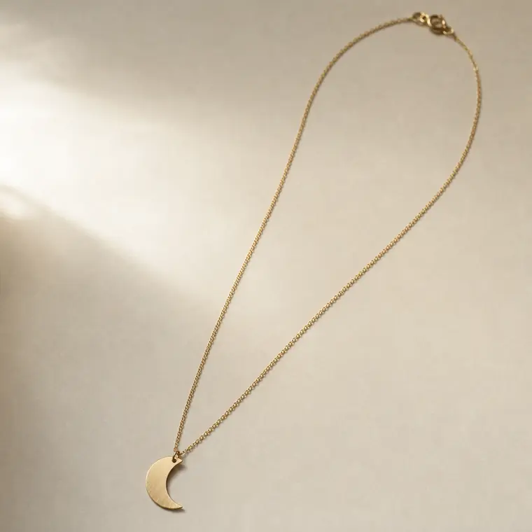 A necklace with a single flat crescent-moon pendant in brushed pale gold metal on a fine chain, laid out straight.