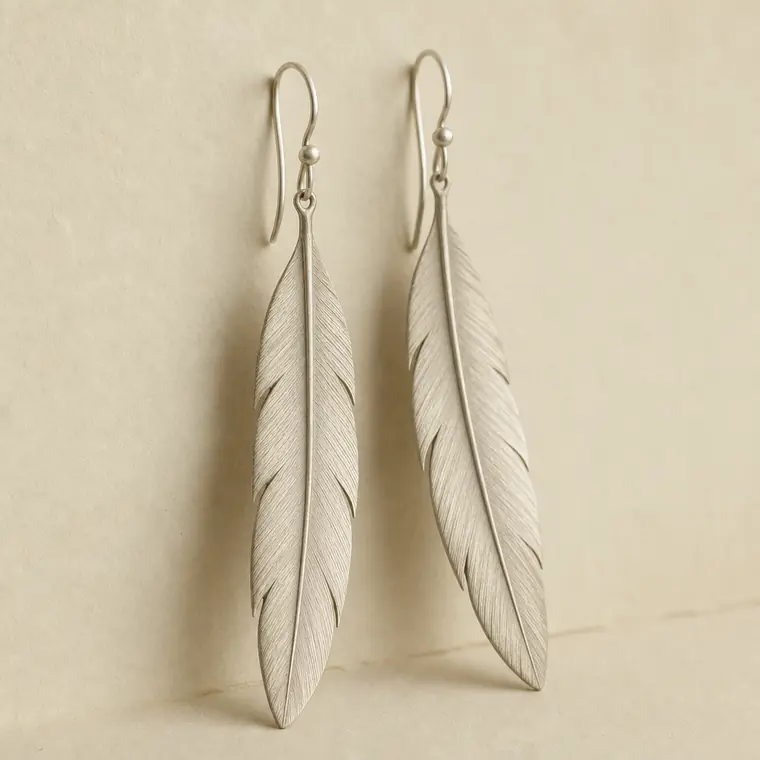 A pair of long feather-shaped drop earrings in brushed pale metal, hanging side by side against ivory paper.